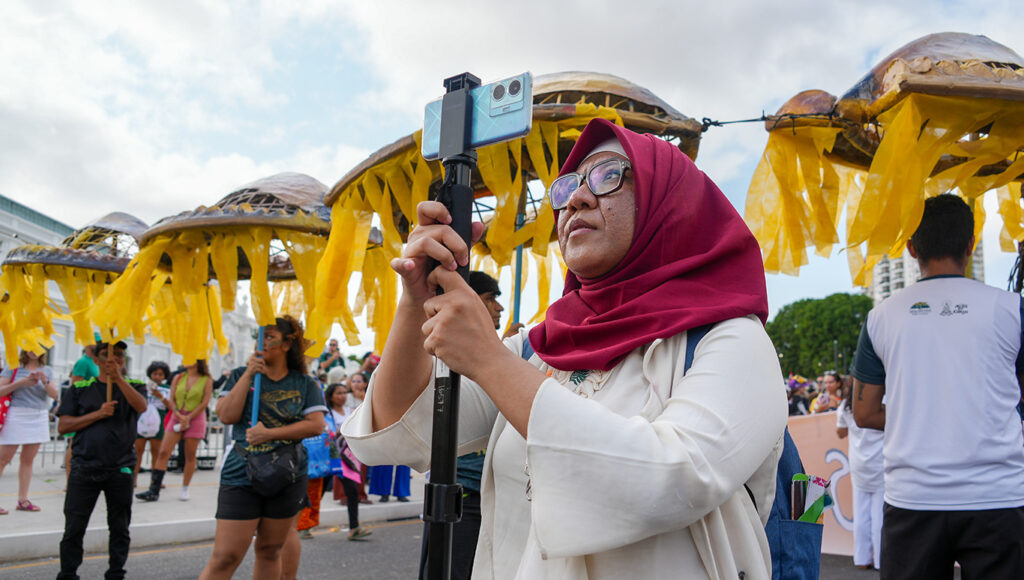 A Climate Change Media Partnership (CCMP) Reporting Fellow documents a march in Belém, Brazil, at COP30. 2026/01/jm_DSC02561cc.jpg